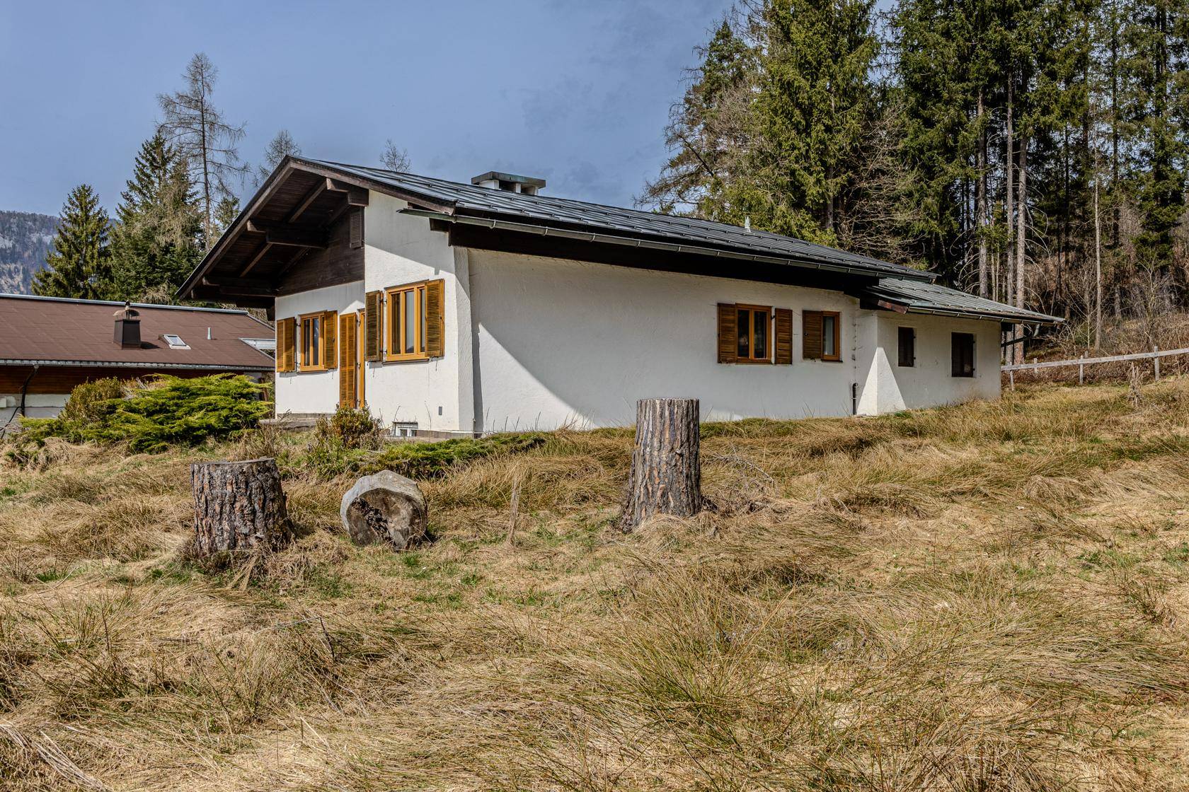 Einfaches weißes Haus mit braunen Fensterläden und Satteldach, umgeben von trockenem Gras und Bäumen.