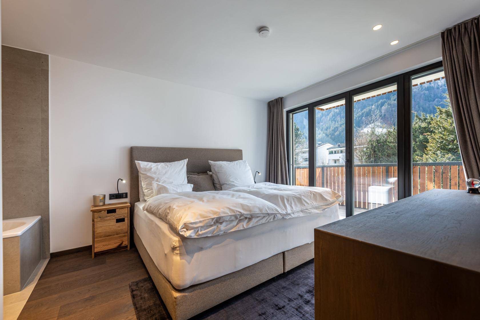 Helles Schlafzimmer mit Doppelbett, Holzboden und Zugang zum Balkon mit Bergblick.