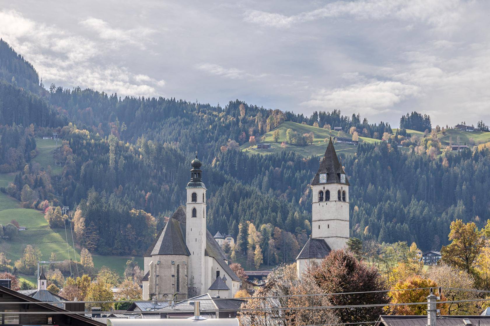 Malerische Ansicht einer Stadt mit zwei Kirchen vor einer bewaldeten Berglandschaft im Herbst.
