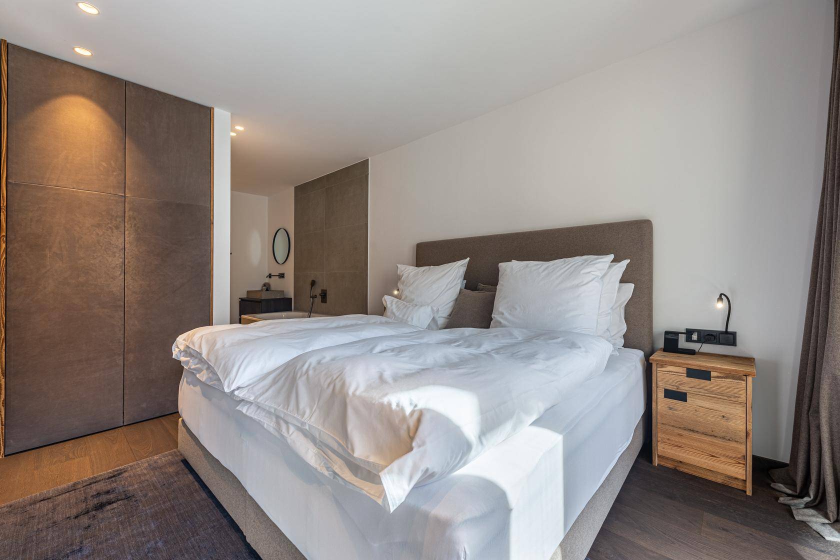 Elegantes Schlafzimmer mit Doppelbett, Holzboden und integriertem Badezimmerbereich.