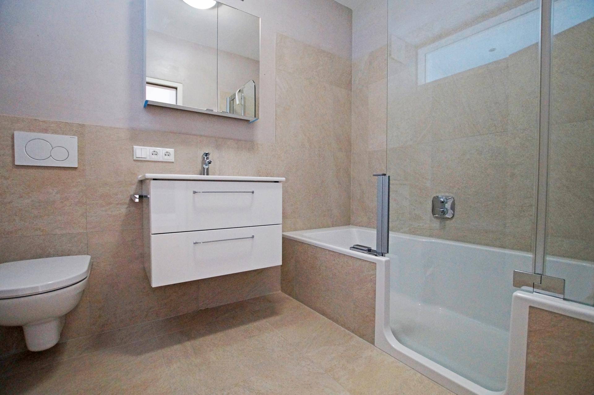 Modernes Badezimmer mit beigefarbenen Fliesen, Badewanne mit Duschabtrennung und wandhängendem WC.