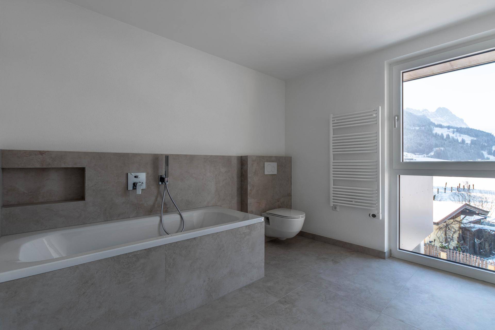 Modernes Badezimmer mit Badewanne, Wand-WC, beheiztem Handtuchhalter und Fenster mit Bergblick.