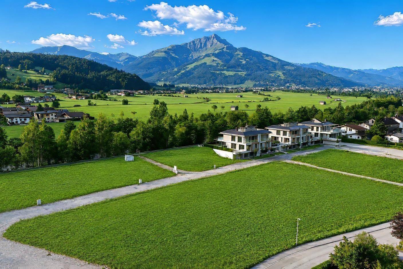 Moderne Wohnanlage mit mehreren Häusern in grüner Umgebung und beeindruckendem Bergblick unter strahlendem Himmel.