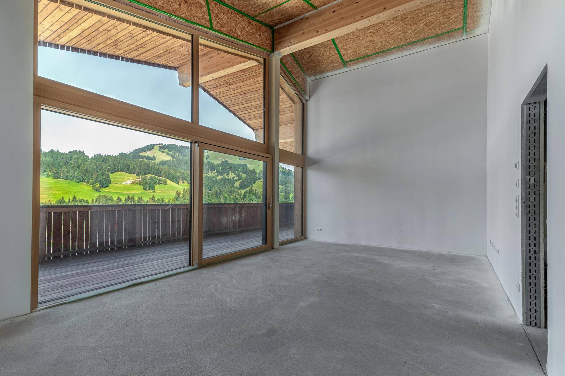 Unfertiger Innenraum mit großen Panoramafenstern, die einen weiten Blick auf die grüne Bergwelt bieten.
