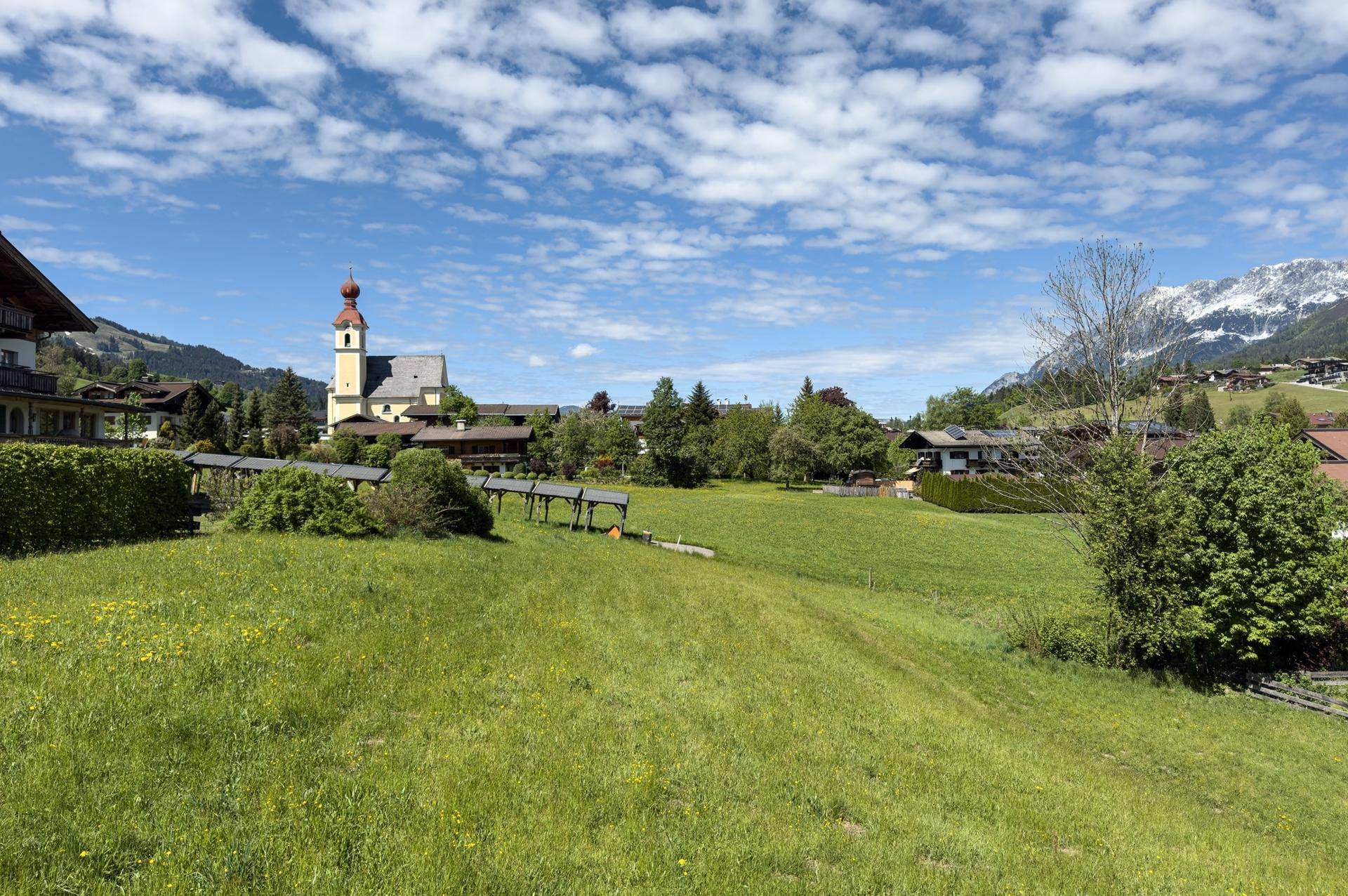 Weitläufige grüne Landschaft mit Blick auf ein charmantes Dorf, eine Kirche und schneebedeckte Berge im Hintergrund.