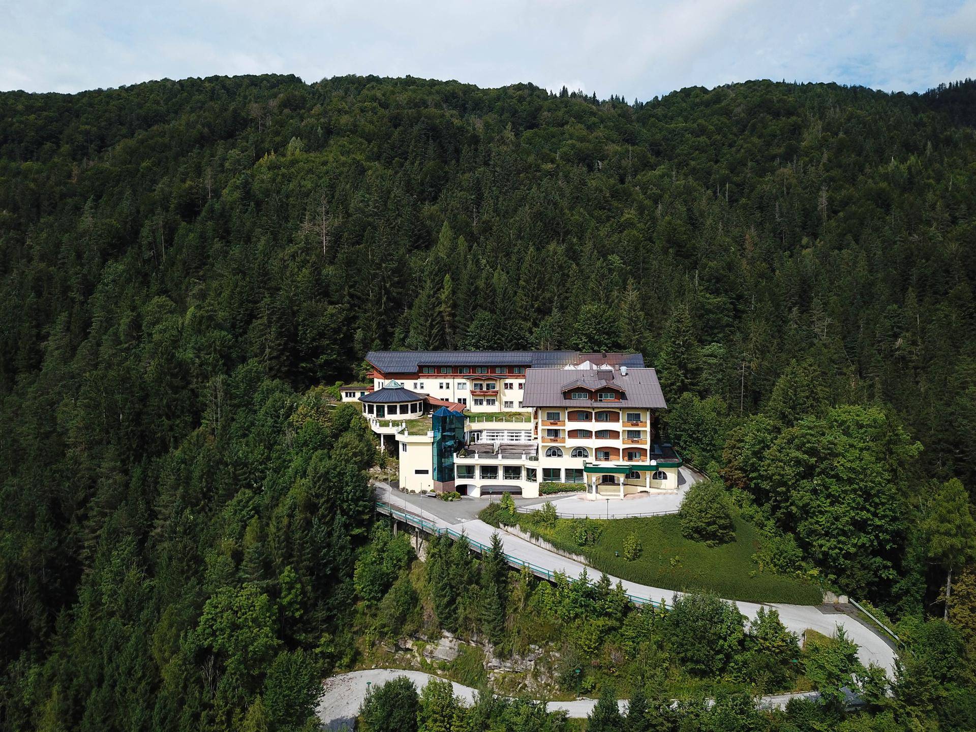 Luftaufnahme eines weitläufigen Gebäudes, eingebettet in einen bewaldeten Berghang mit einer Serpentinenstraße.