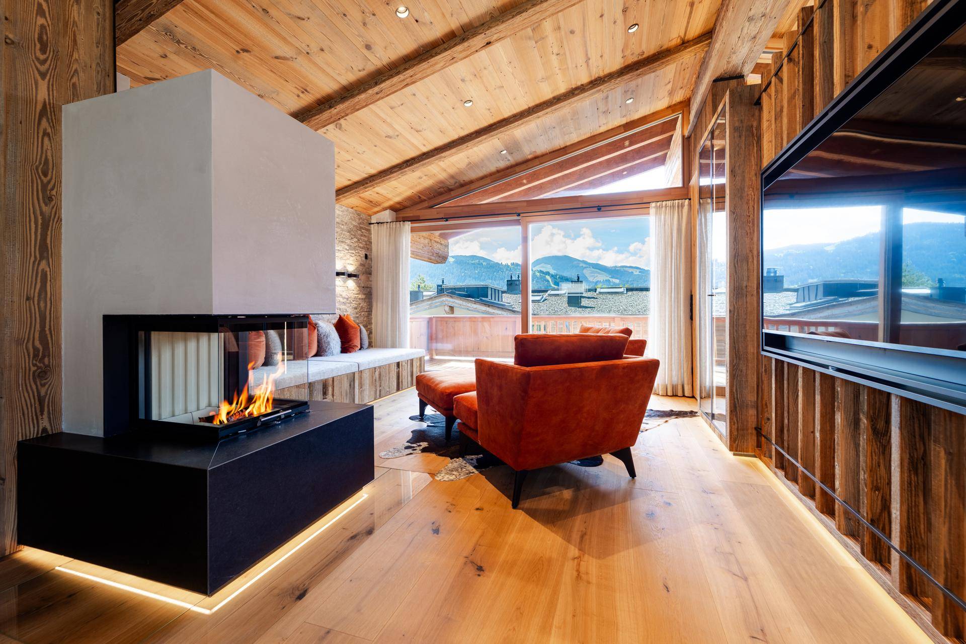 Gemütliches Wohnzimmer mit modernem Kamin, Holzbalkendecke und weitem Blick auf die Berglandschaft.