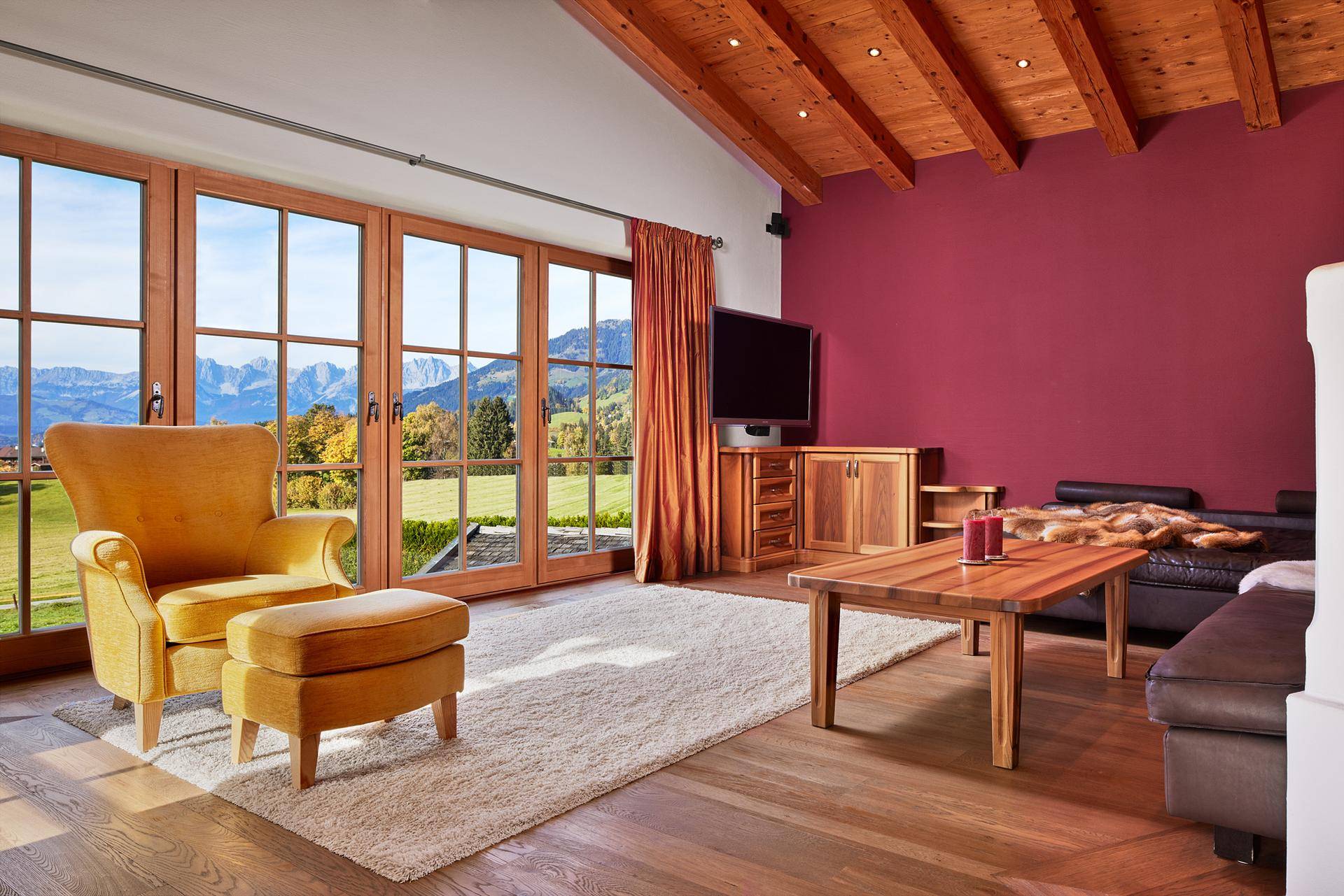 Helles Wohnzimmer mit roter Akzentwand, Holzboden und weitem Blick auf die Berge und grüne Landschaft.
