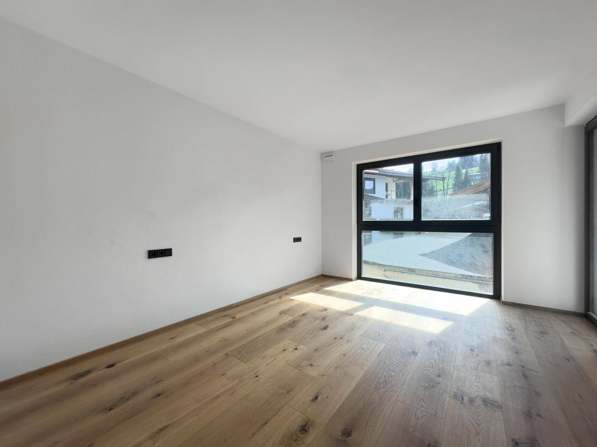 Lichtdurchflutetes Zimmer mit Holzboden und großem Fenster, das einen Blick ins Grüne ermöglicht.