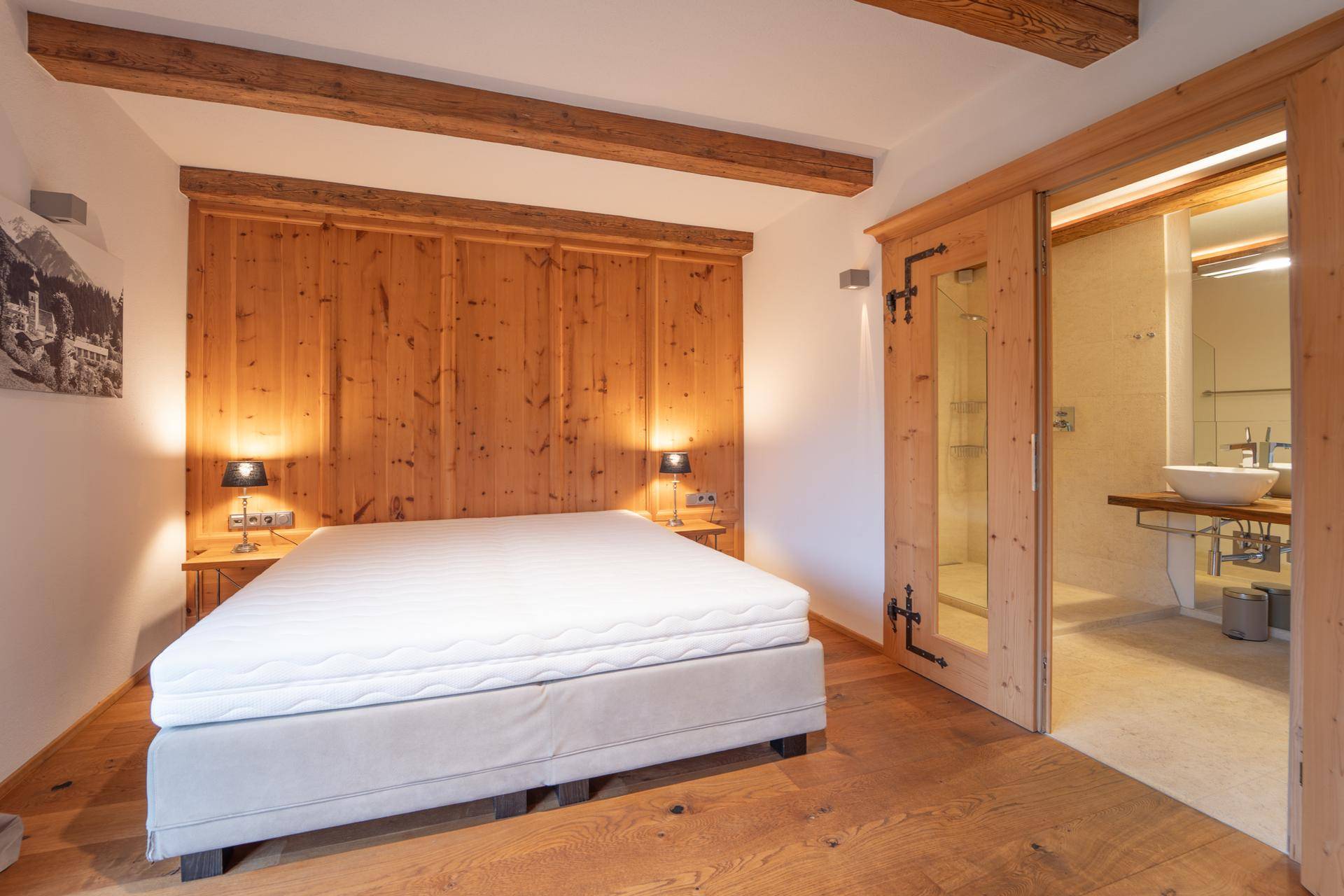 Helles Schlafzimmer mit Holzwand, Holzbalken und direktem Zugang zum Bad.