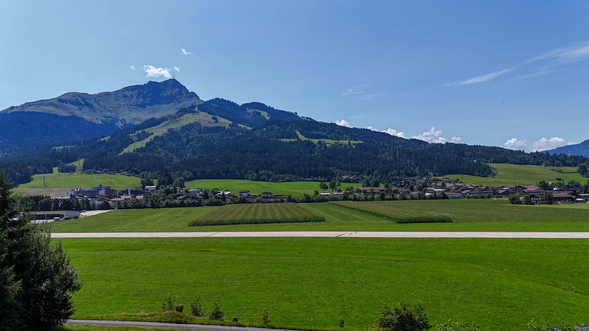 Panoramablick auf grüne Felder, eine Landebahn und majestätische Berge im Hintergrund.