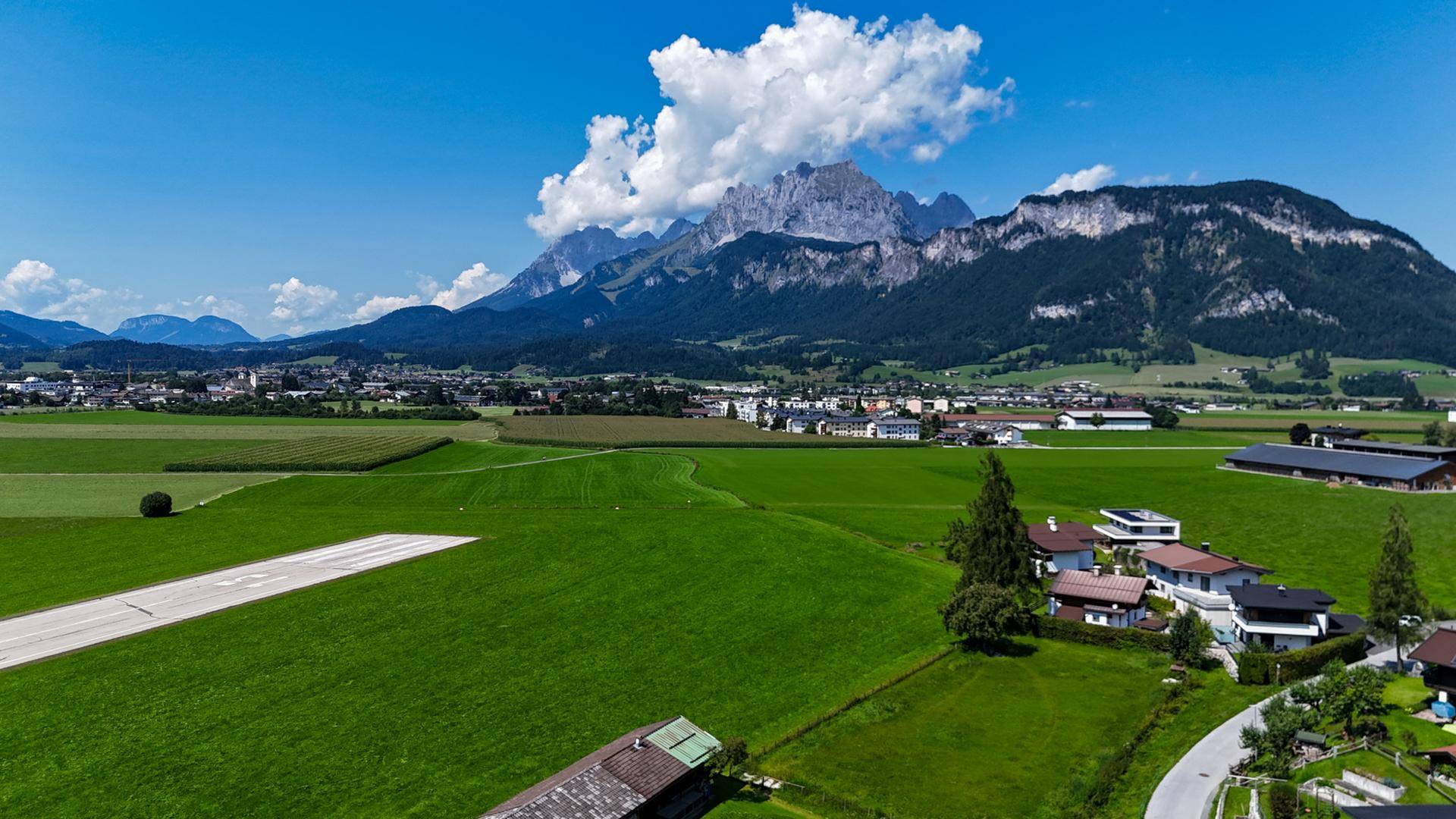 Weitläufige Landschaft mit einem Dorf, Bergen und einem Flugfeld unter blauem Himmel.
