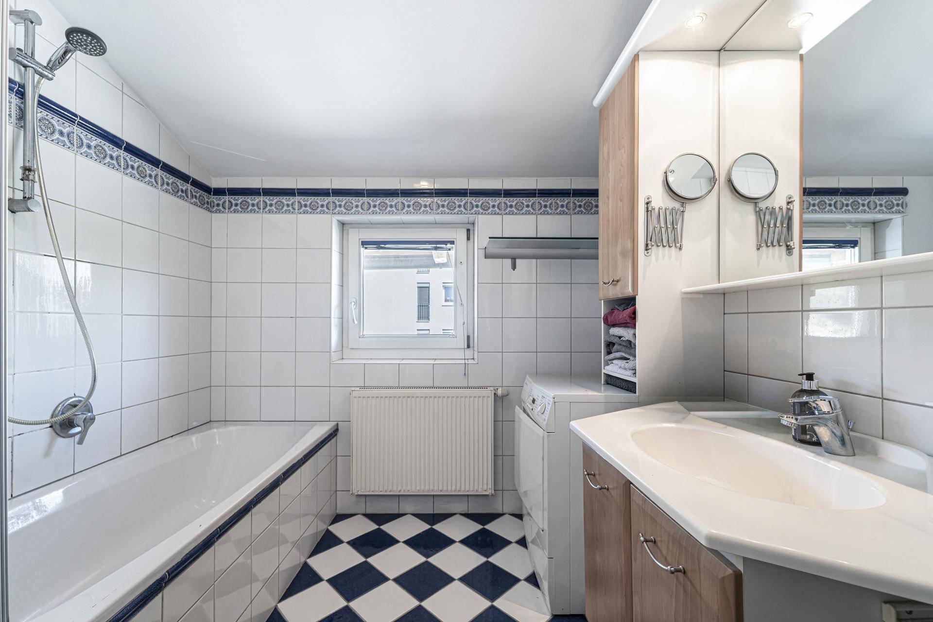 Badezimmer mit Badewanne, Dusche, Waschbecken, Waschmaschine und Fenster in weiß-blauer Gestaltung.