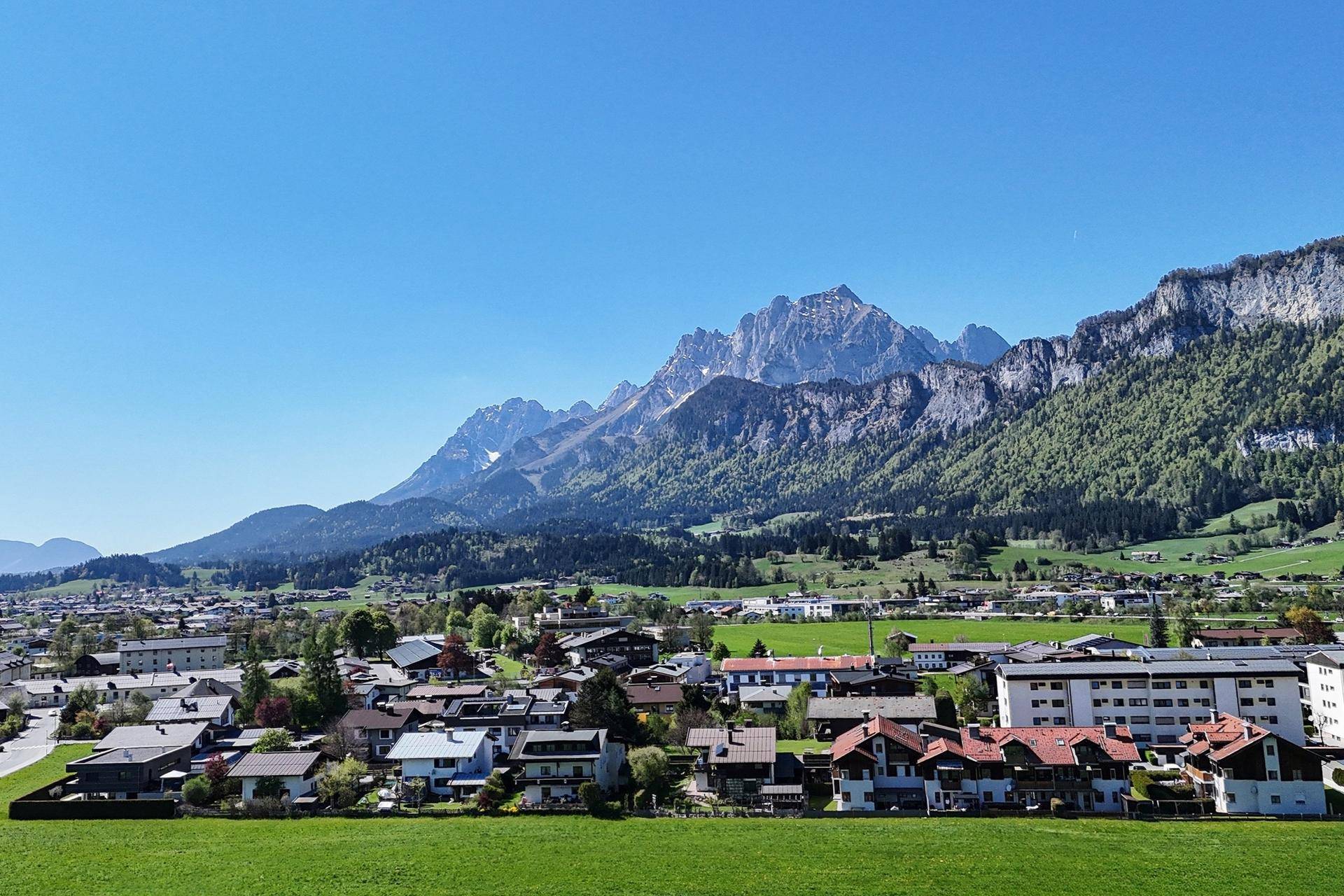 Panoramablick auf ein malerisches Dorf mit grünen Wiesen und majestätischen Bergen im Hintergrund.