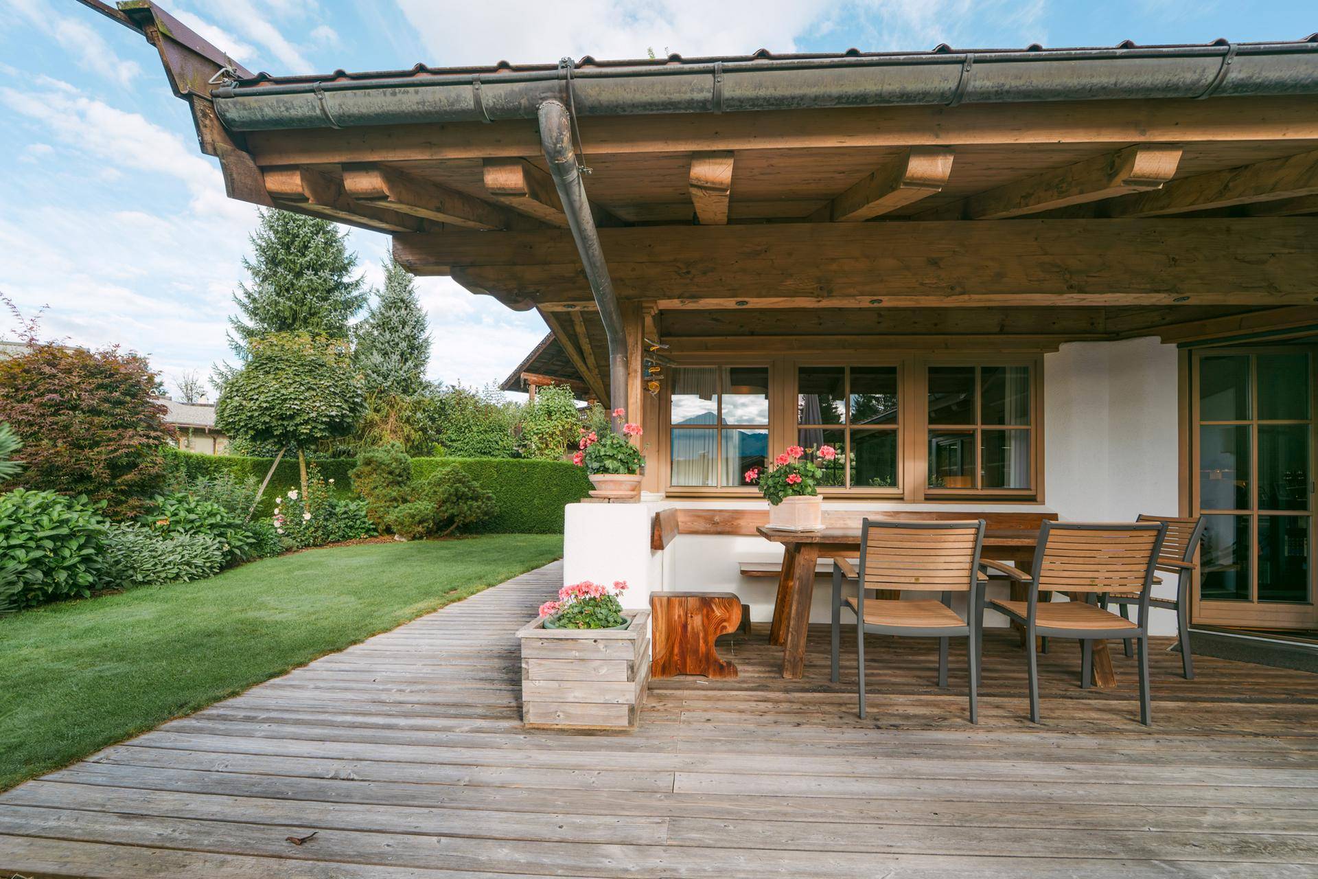Gemütliche Holzterrasse mit Esstisch und Stühlen, umgeben von üppigem Grün und Blumen.