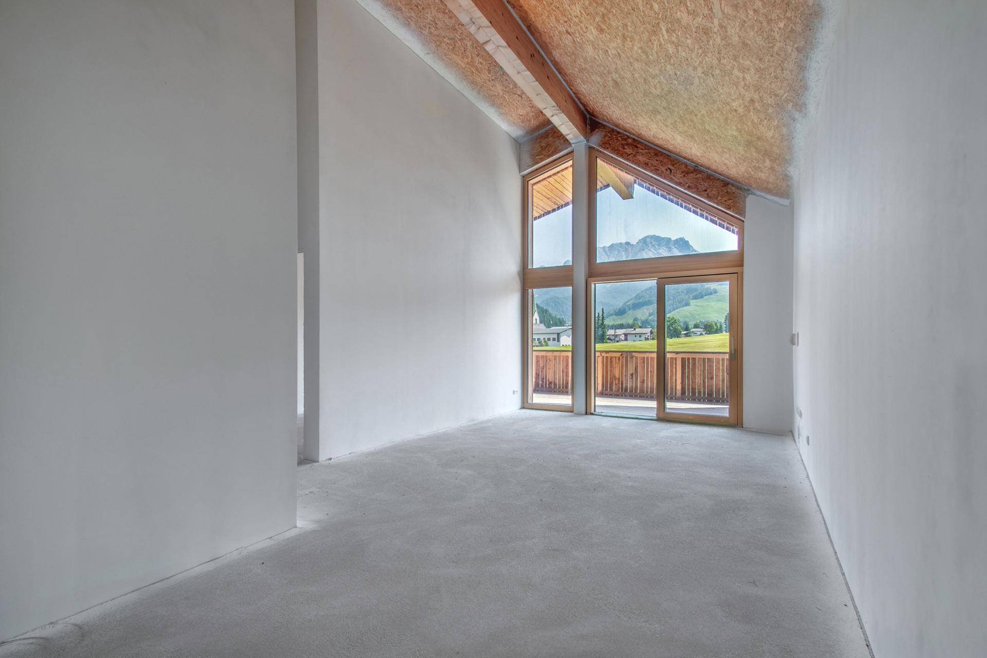 Leerer Raum im Rohbau mit hohen, gewölbten Holzdecken, großen Fenstern und Bergblick.