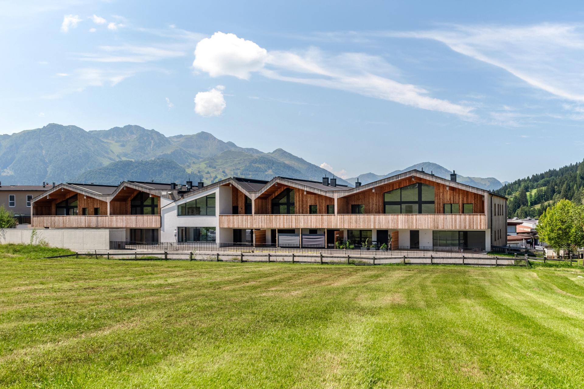 Reihe moderner Gebäude mit Holzfassaden und großen Fenstern vor alpiner Bergkulisse.