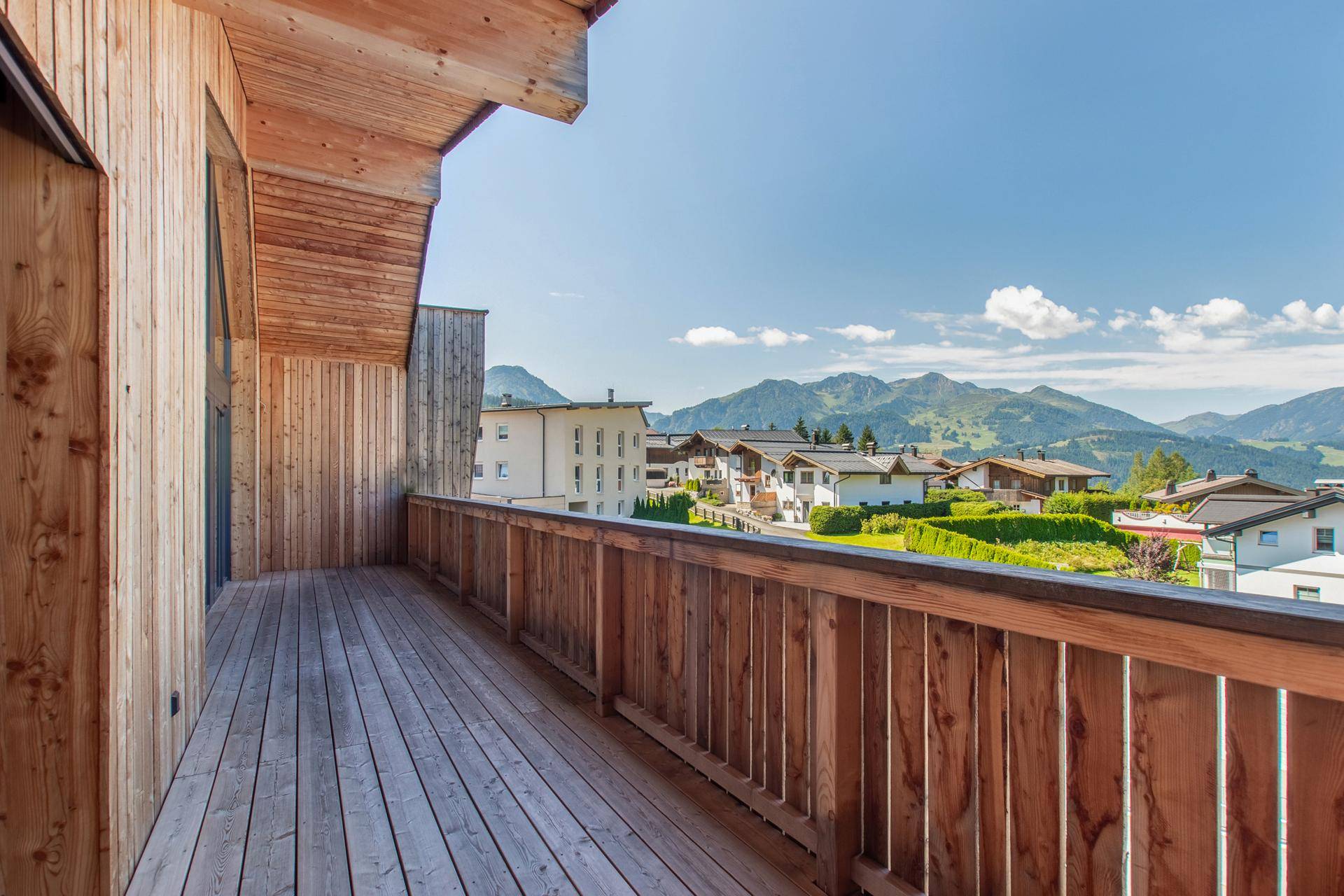 Geräumiger Holzbalkon mit Blick auf ein Dorf und die umliegende Berglandschaft unter blauem Himmel.