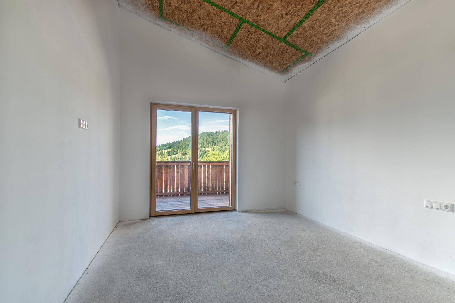 Unfertiger Raum mit Betonboden und Zugang zu einem Holzbalkon mit Blick ins Grüne.