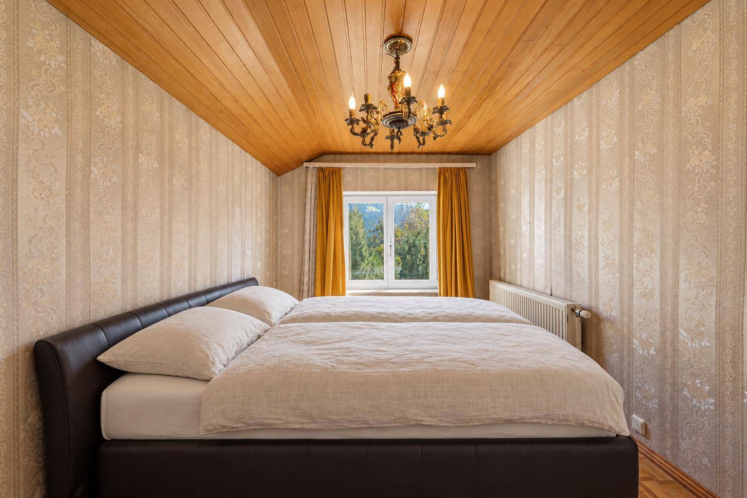 Schlafzimmer mit Holzdecke, gemusterter Tapete und Fenster mit Blick ins Grüne.