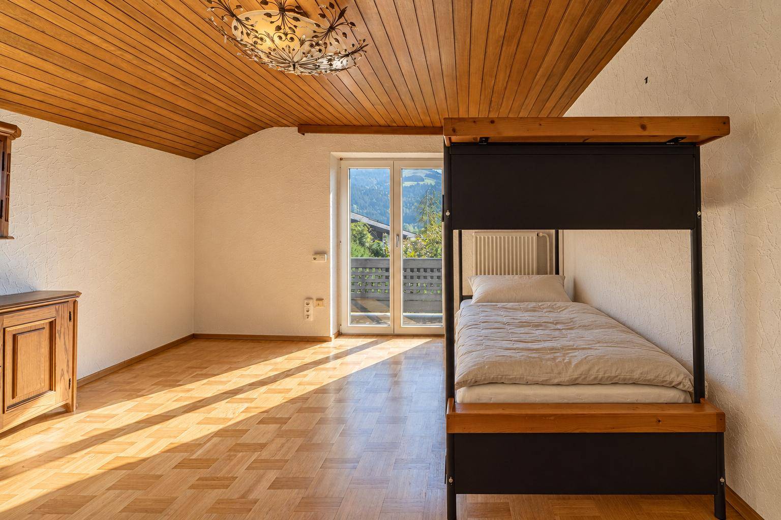 Helles Schlafzimmer mit Parkettboden, Holzdecke und Zugang zum Balkon mit Bergblick.