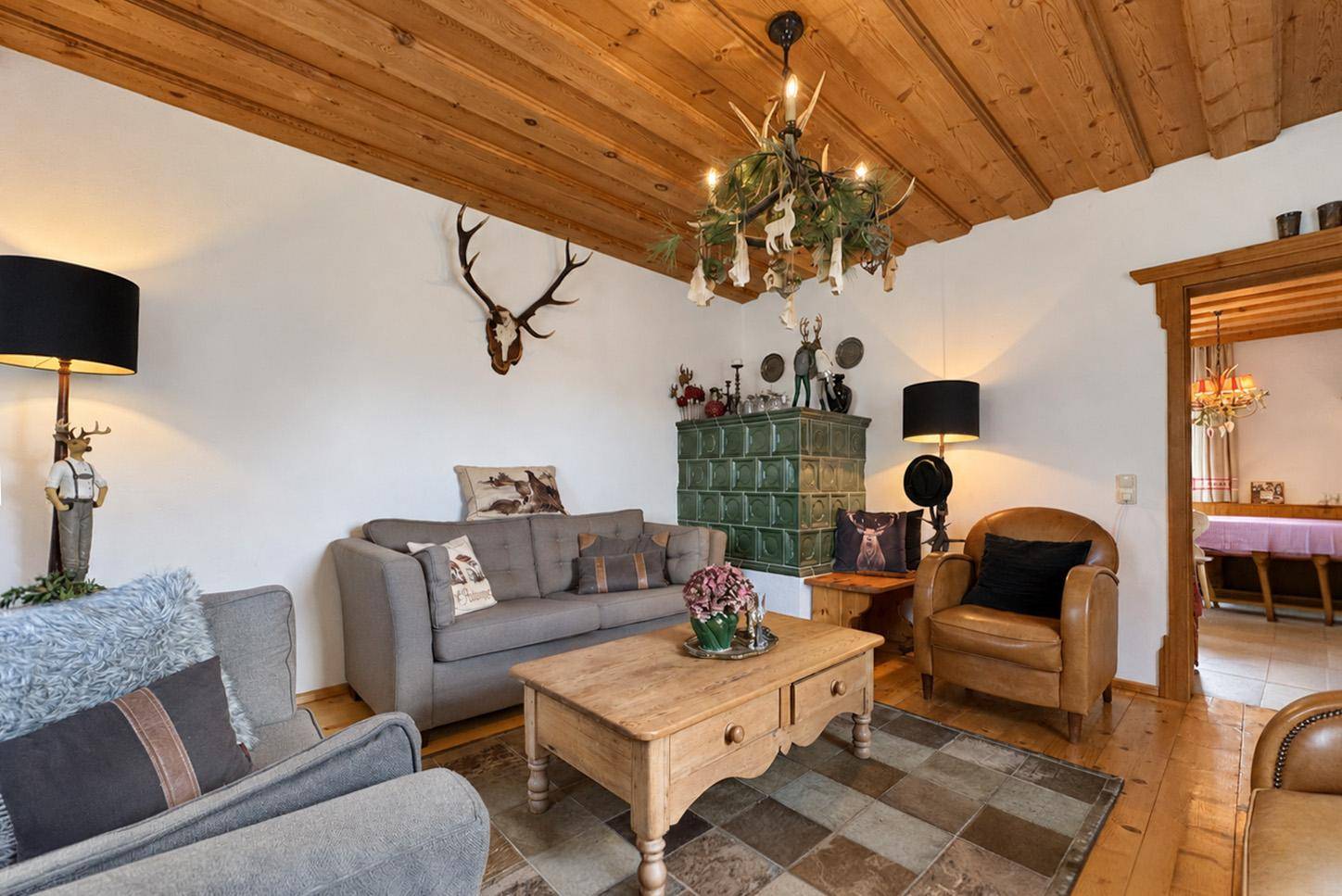 Gemütliches Wohnzimmer im alpinen Stil mit Kachelofen, Holzdecke und Hirschgeweih-Dekoration.
