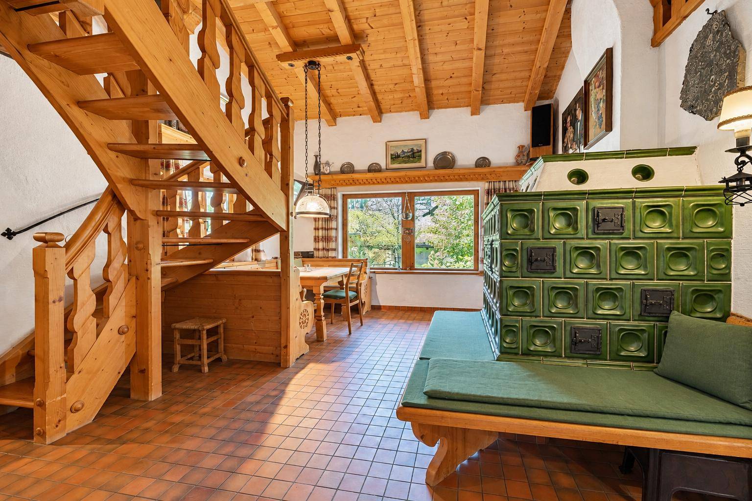 Traditioneller Wohnbereich mit grünem Kachelofen, Holztreppe und rustikalen Fliesen.