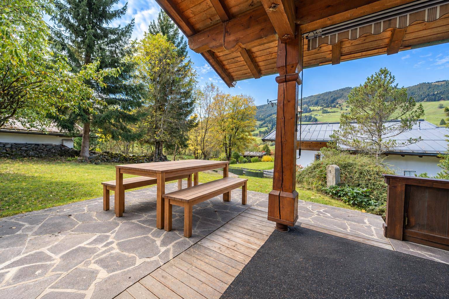 Gemütliche Terrasse mit Holztisch und Bänken, umgeben von grüner Natur und Bergblick.