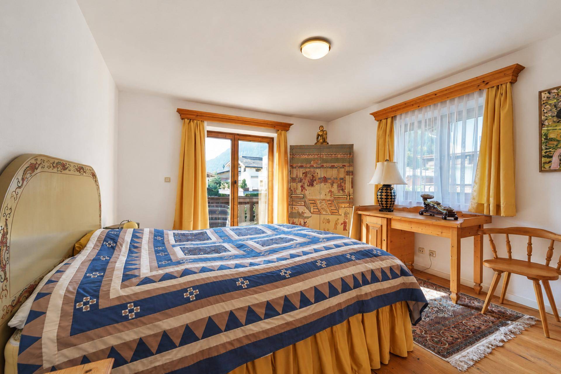 Helles Schlafzimmer mit Holzbett, Schreibtisch und Balkonzugang mit Bergblick.