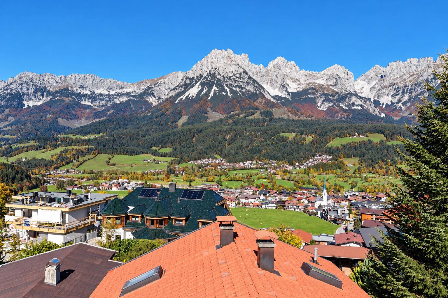 Panoramablick über ein malerisches Dorf mit grünen Feldern und imposanten Bergen im Hintergrund.