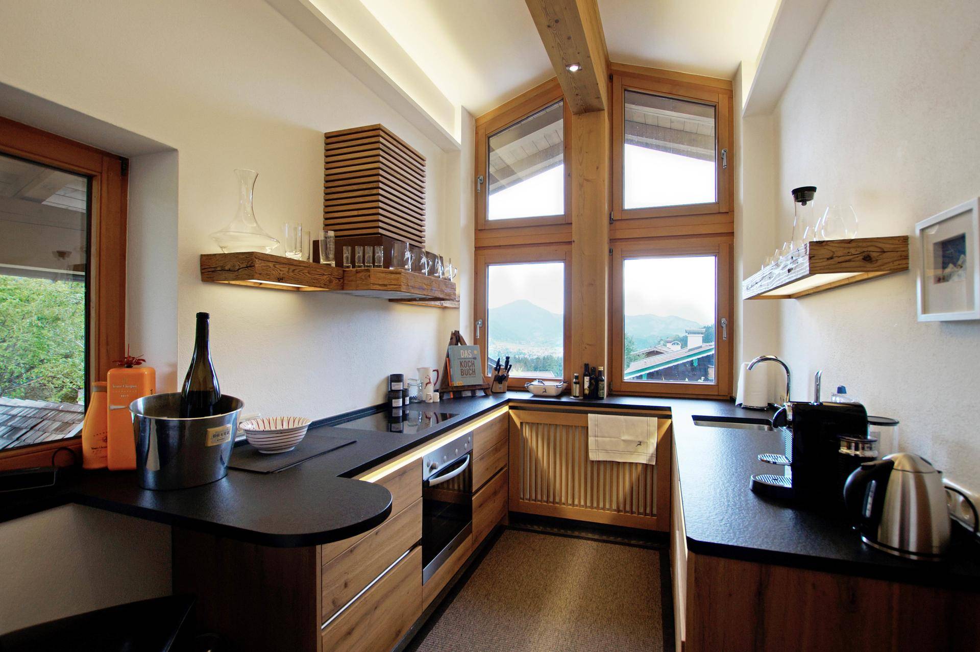 Moderne Küche mit Holzelementen, dunkler Arbeitsplatte und großen Fenstern mit Bergblick.