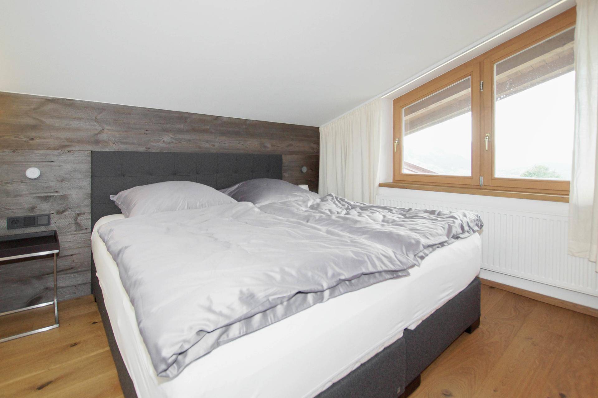 Helles Schlafzimmer mit Holzwand, Doppelbett und großem Fenster mit Blick ins Grüne.