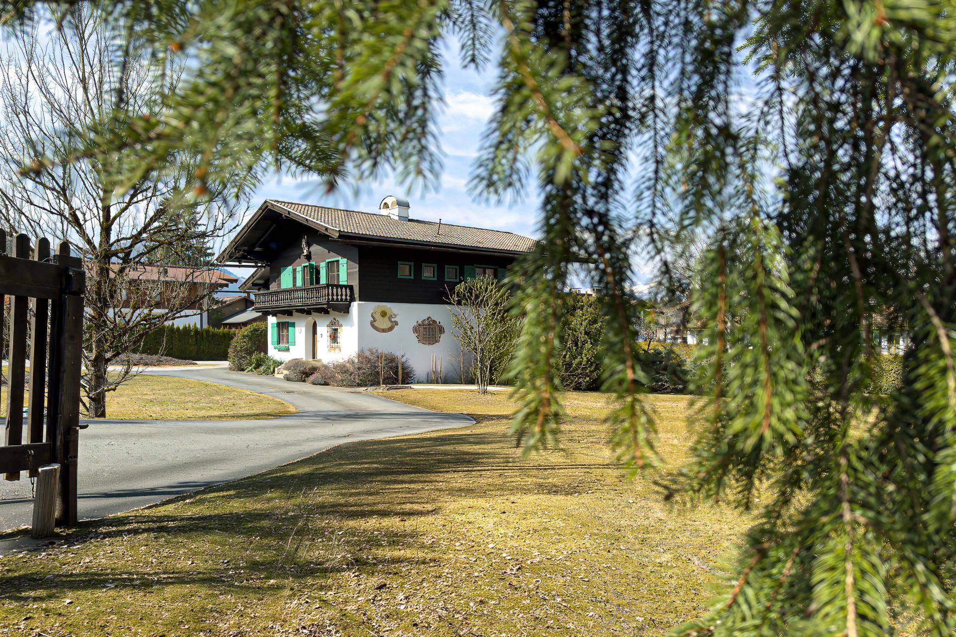 Charmantes Alpenhaus mit grünen Fensterläden, umgeben von Bäumen und einem gepflegten Rasen, mit Bergblick im Hintergrund.