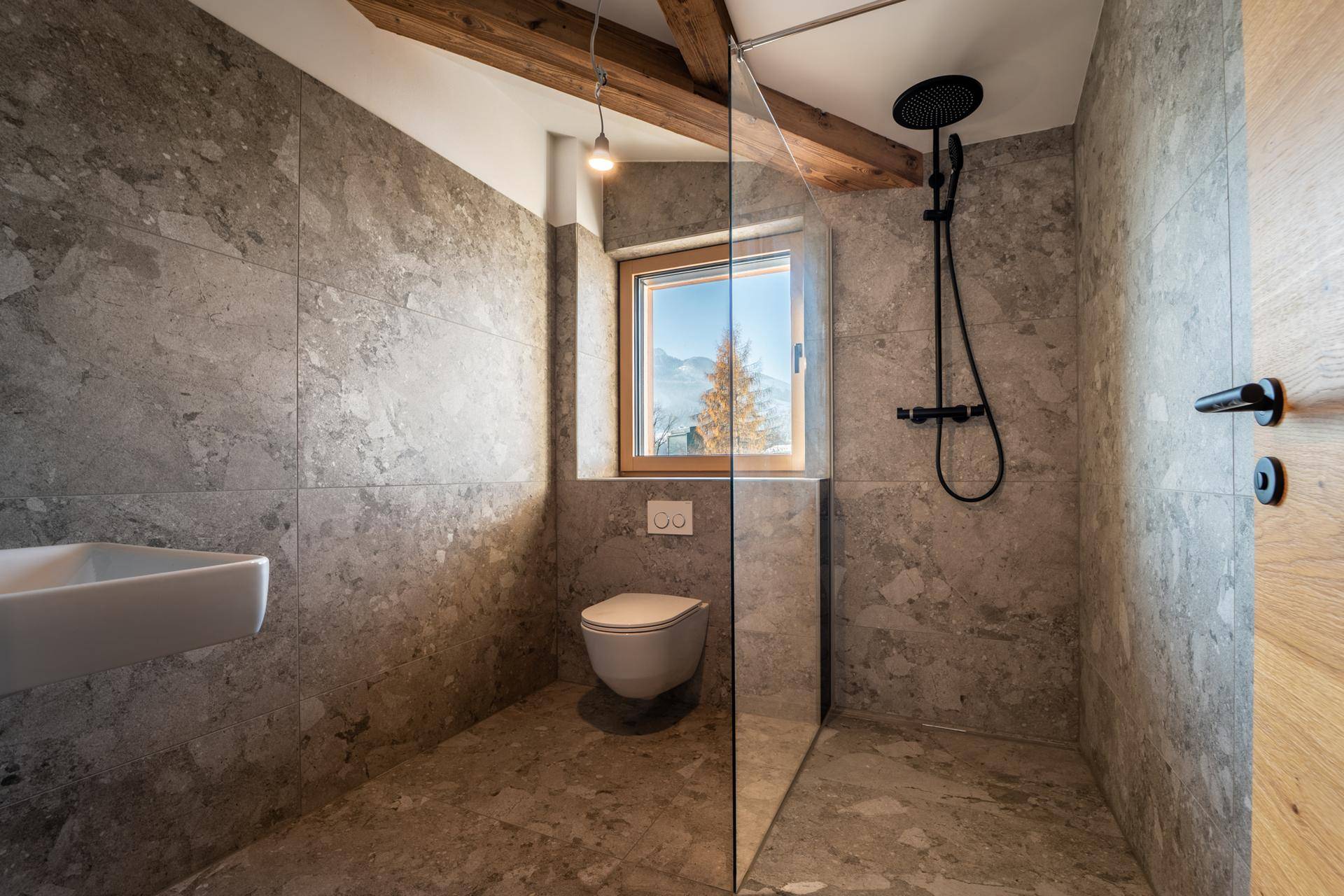 Zeitgemäßes Badezimmer mit ebenerdiger Dusche, Wand-WC und Fenster, komplett gefliest in Grau.