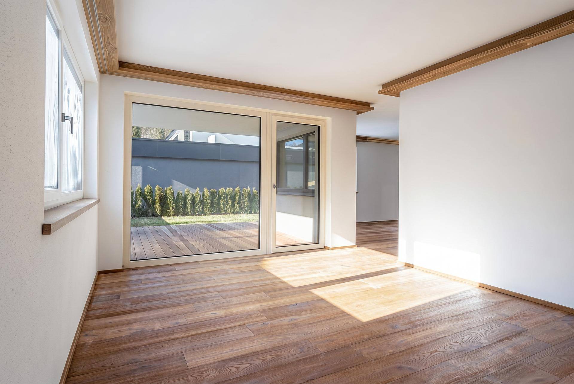 Helles, leeres Wohnzimmer mit Holzboden, sichtbaren Holzbalken und großen Fenstern zum Garten.