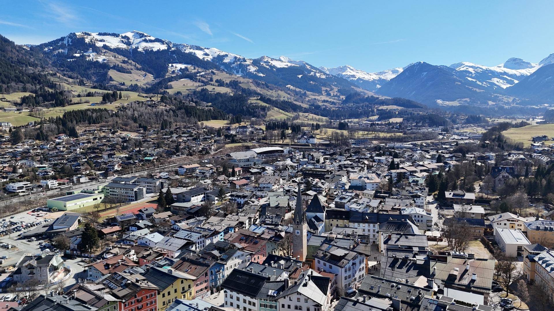Panoramablick auf eine malerische Stadt in den Alpen, umgeben von grünen Hügeln und Bergen.