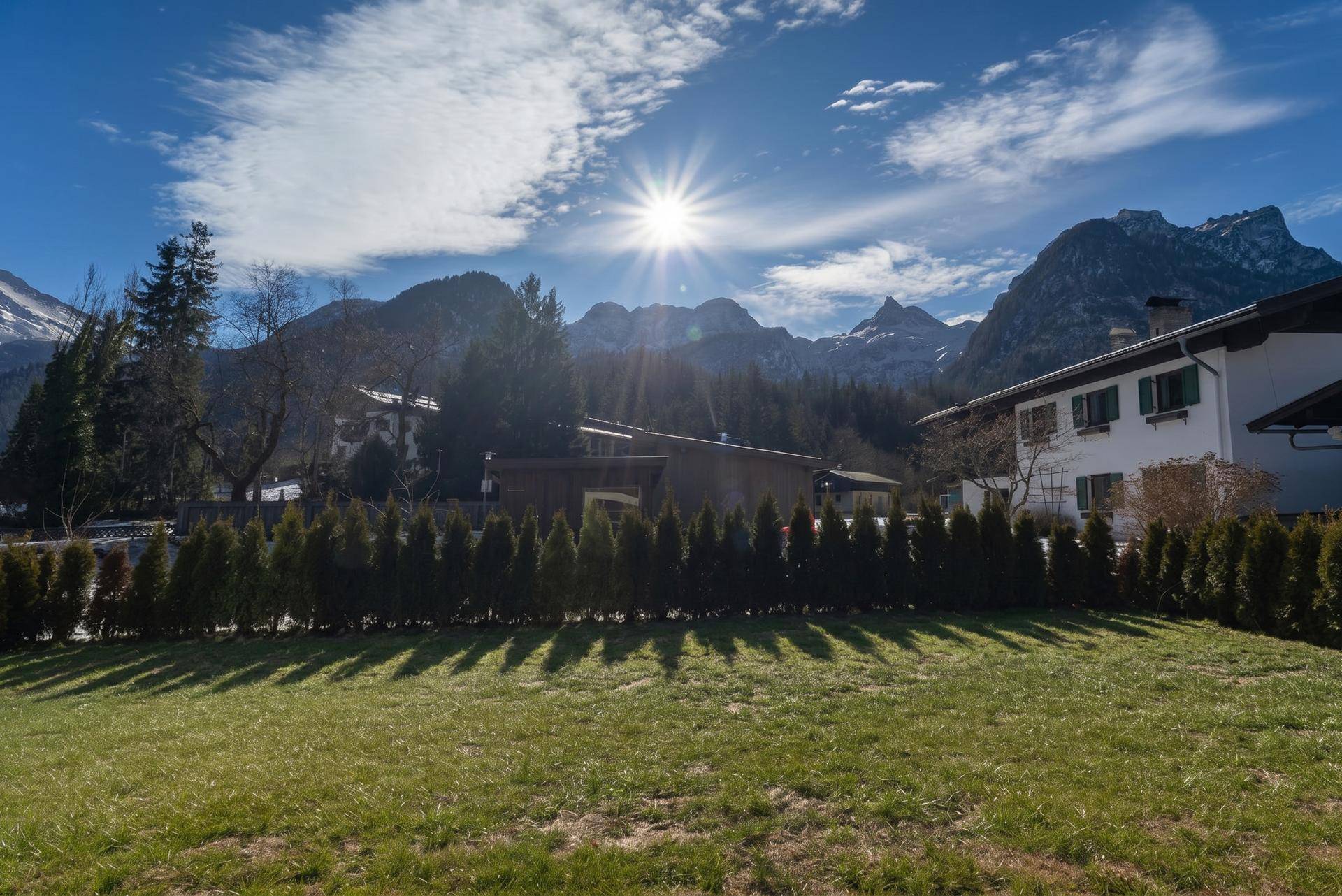 Sonnige Landschaft mit grüner Wiese, Hecke und einem Haus vor einer beeindruckenden Bergkulisse unter blauem Himmel.
