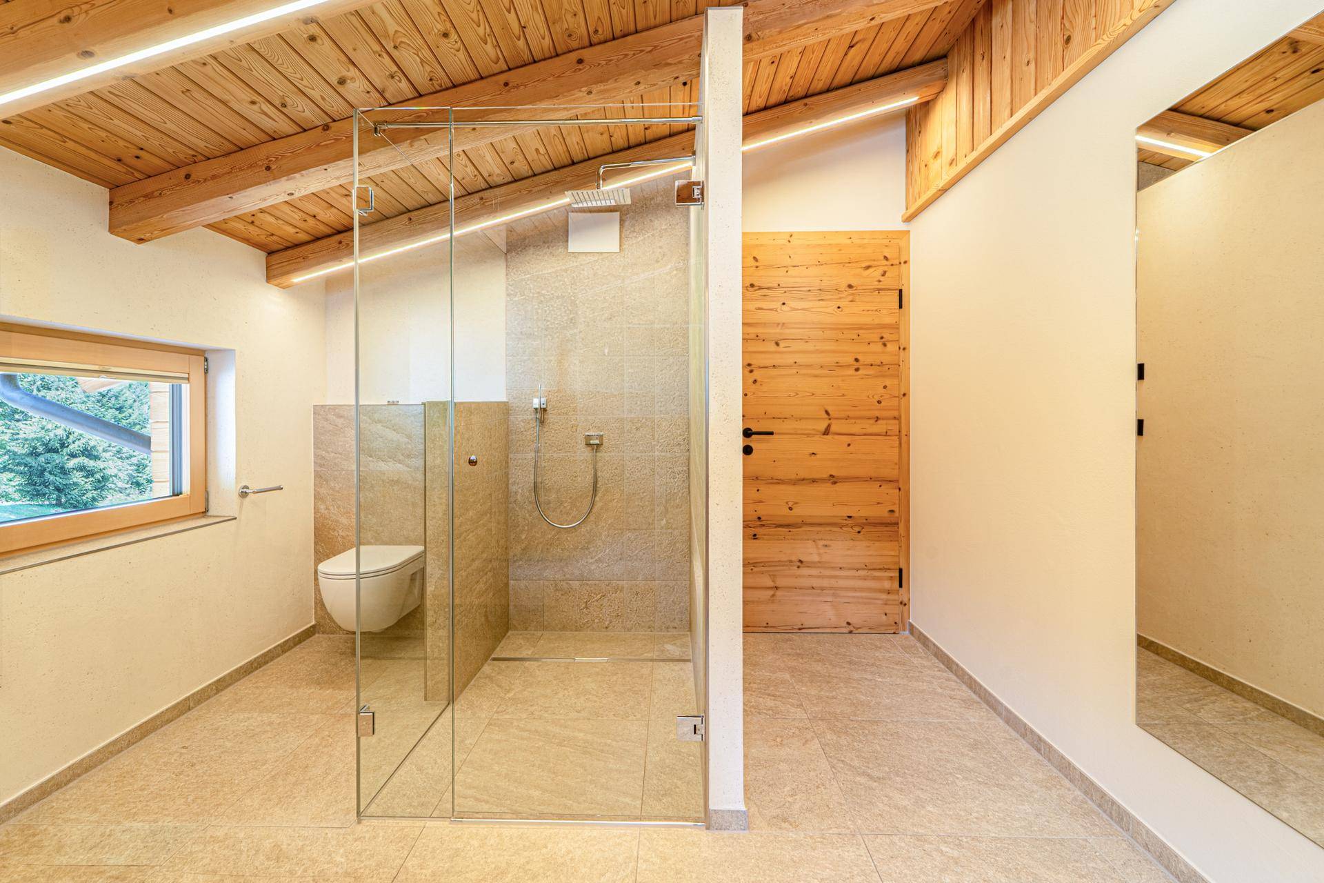 Geräumiges Badezimmer mit begehbarer Dusche, Wand-WC und Holztür, unter einer Holzbalkendecke.