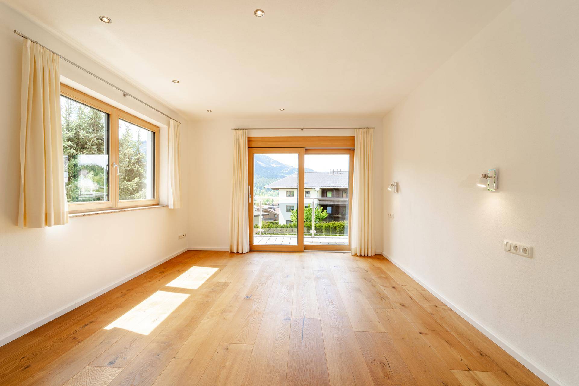 Lichtdurchfluteter Raum mit Holzboden, großen Fenstern und Zugang zu einem Balkon.