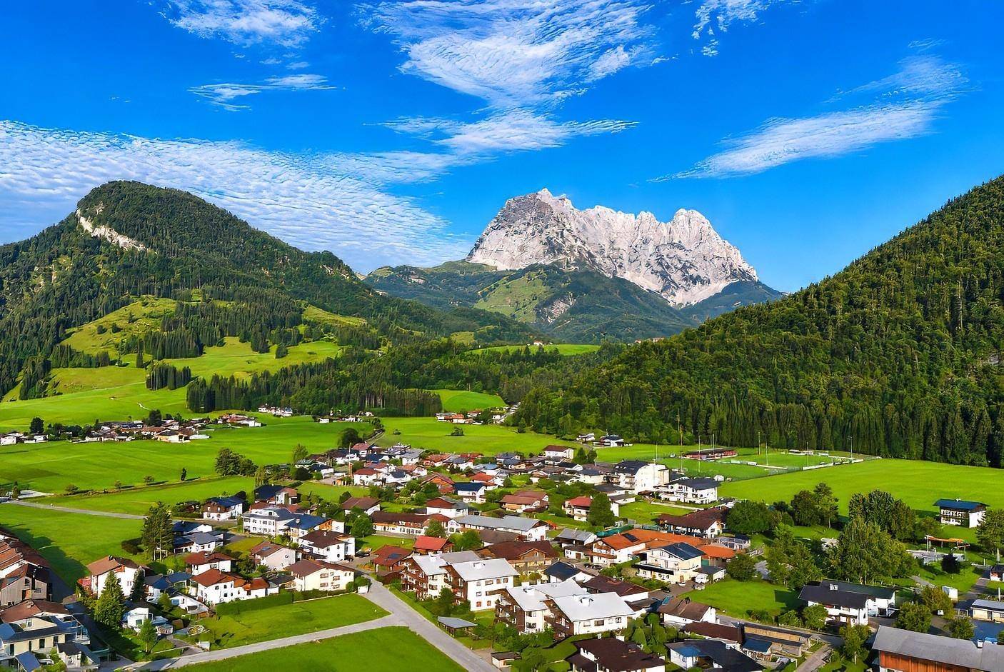 Panoramablick auf ein malerisches Dorf, eingebettet in grüne Hügel mit imposanten Berggipfeln im Hintergrund.
