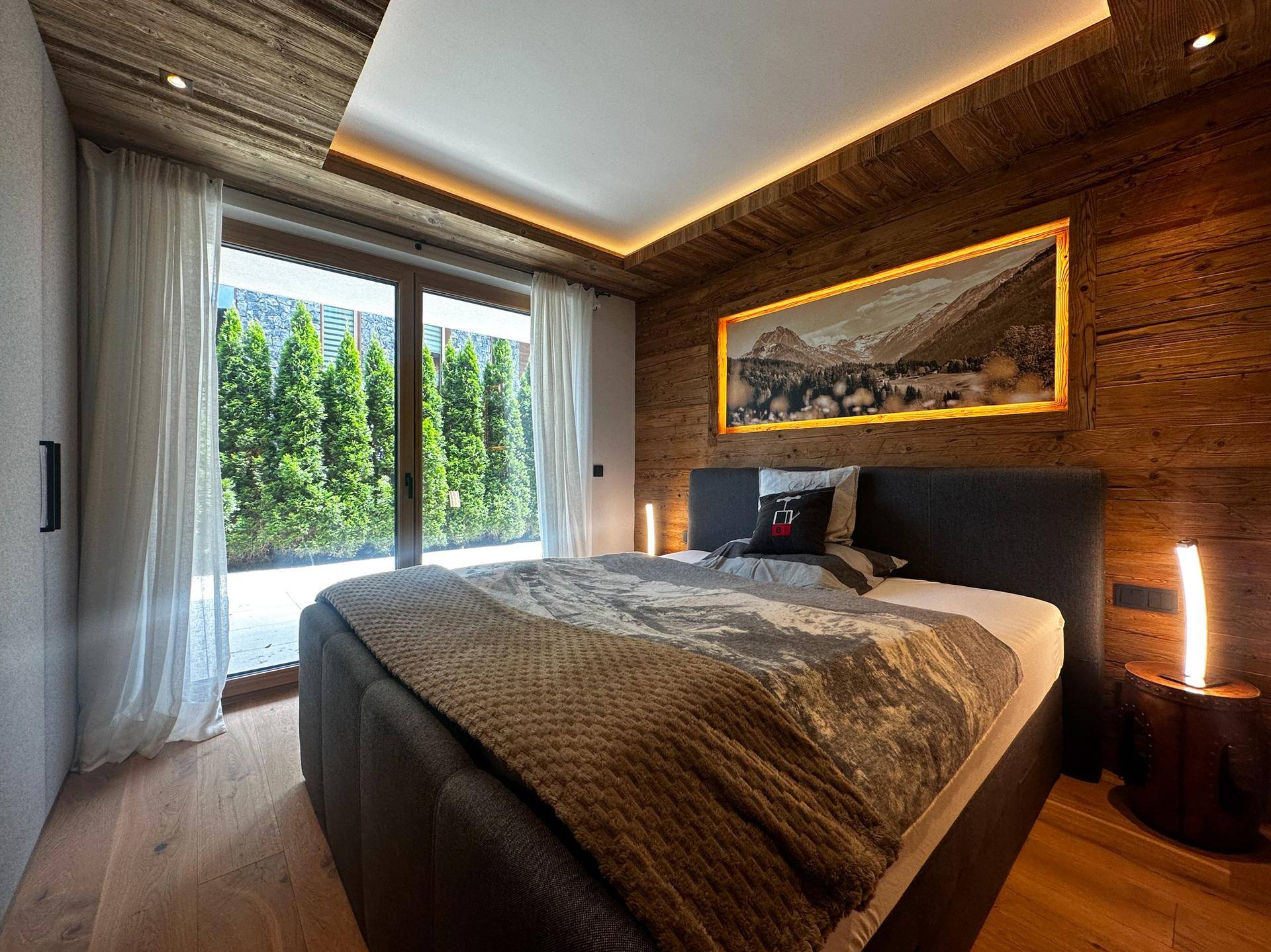 Helles Schlafzimmer mit Holzwänden, großem Fenster und Blick auf die grüne Hecke im Außenbereich.