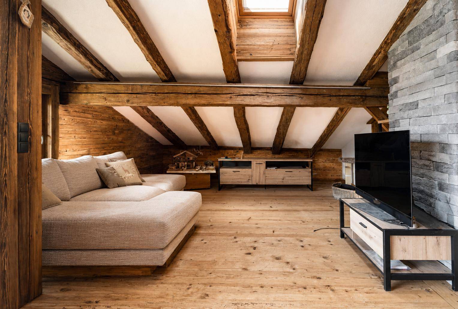 Heller Wohnbereich mit Holzbalken, Holzboden und modernem Sofa unter einem Dachfenster.