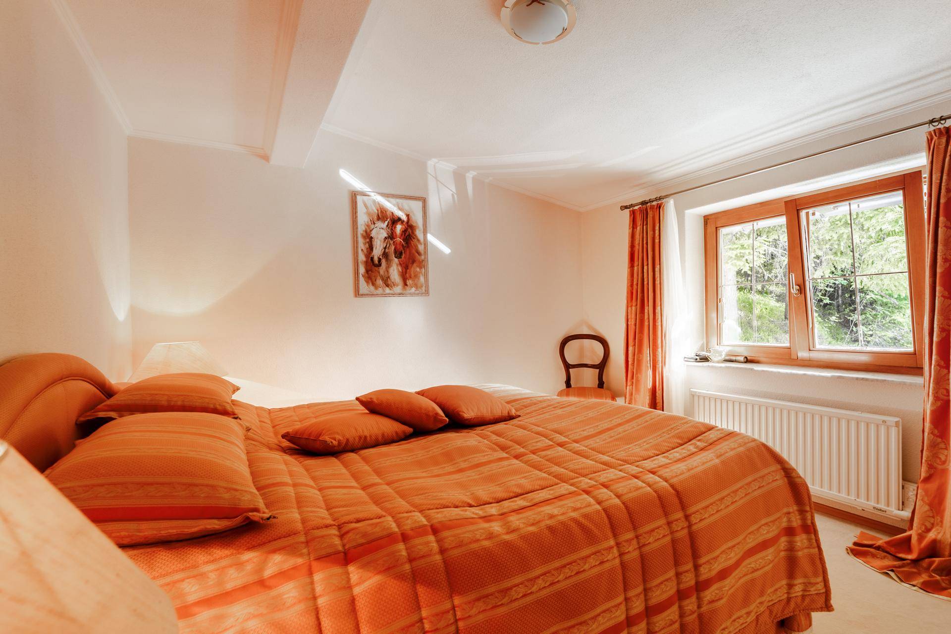 Helles Schlafzimmer mit orangefarbener Bettwäsche und Fenster mit Blick ins Grüne.