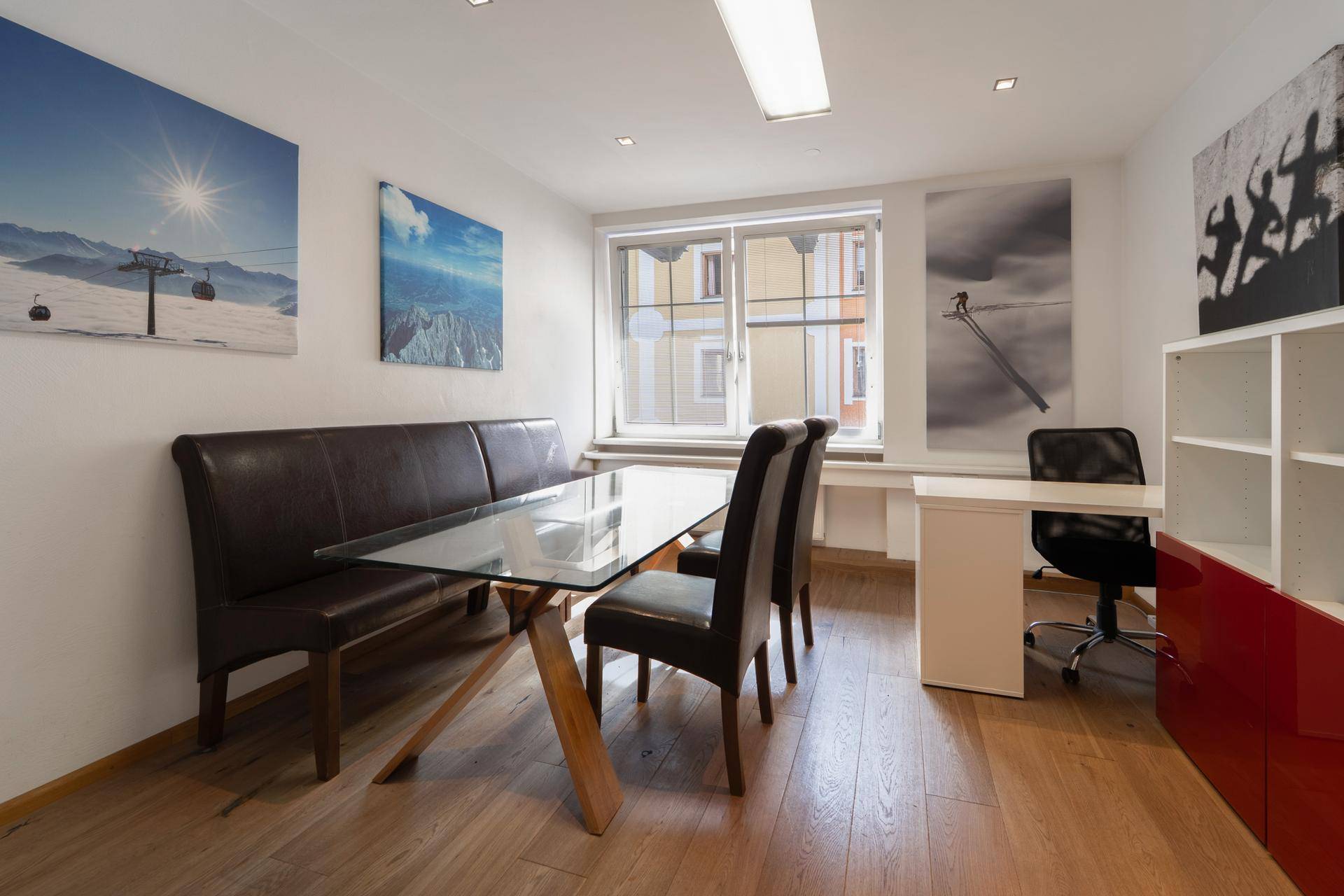 Büro mit Sitzbank, Glastisch, Schreibtisch und Bildern an den Wänden.