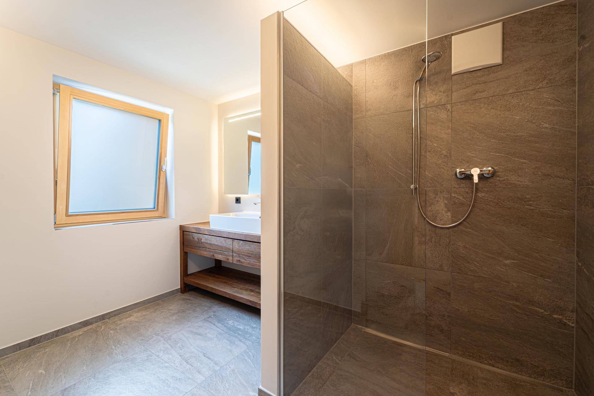 Modernes Badezimmer mit begehbarer Dusche, Waschtisch und Fenster, in neutralen Grau- und Brauntönen.