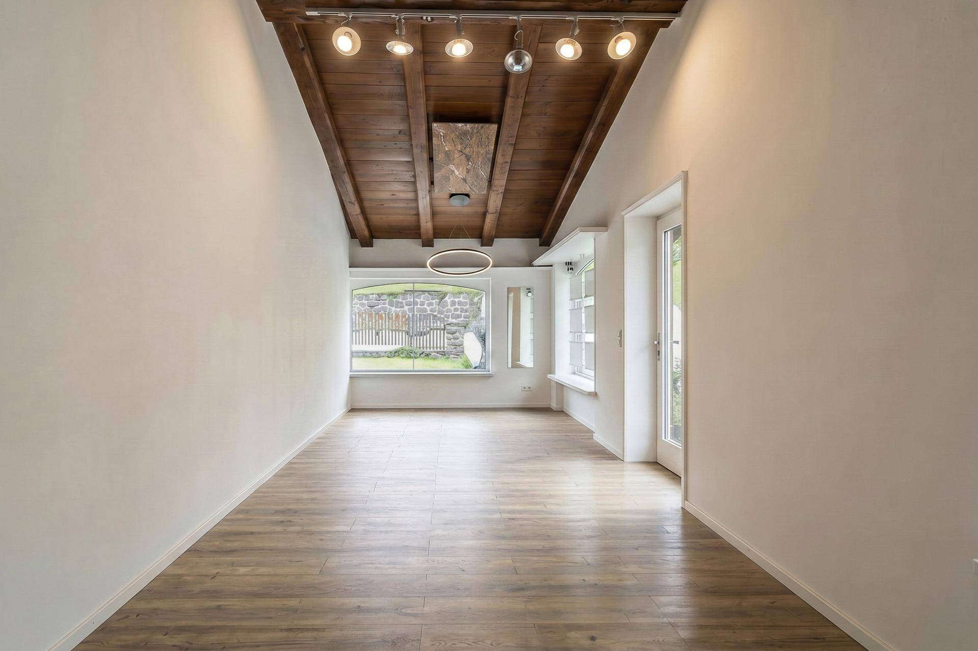Leerer Raum mit Holzbalkendecke, Laminatboden und großen Fenstern, bereit zur individuellen Gestaltung.