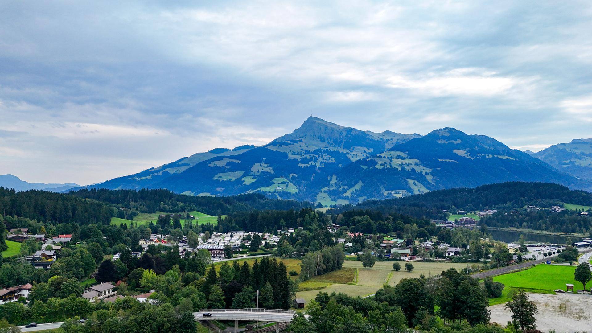 Weitläufige Landschaft mit majestätischen Bergen, Wäldern und einem Dorf am Horizont.