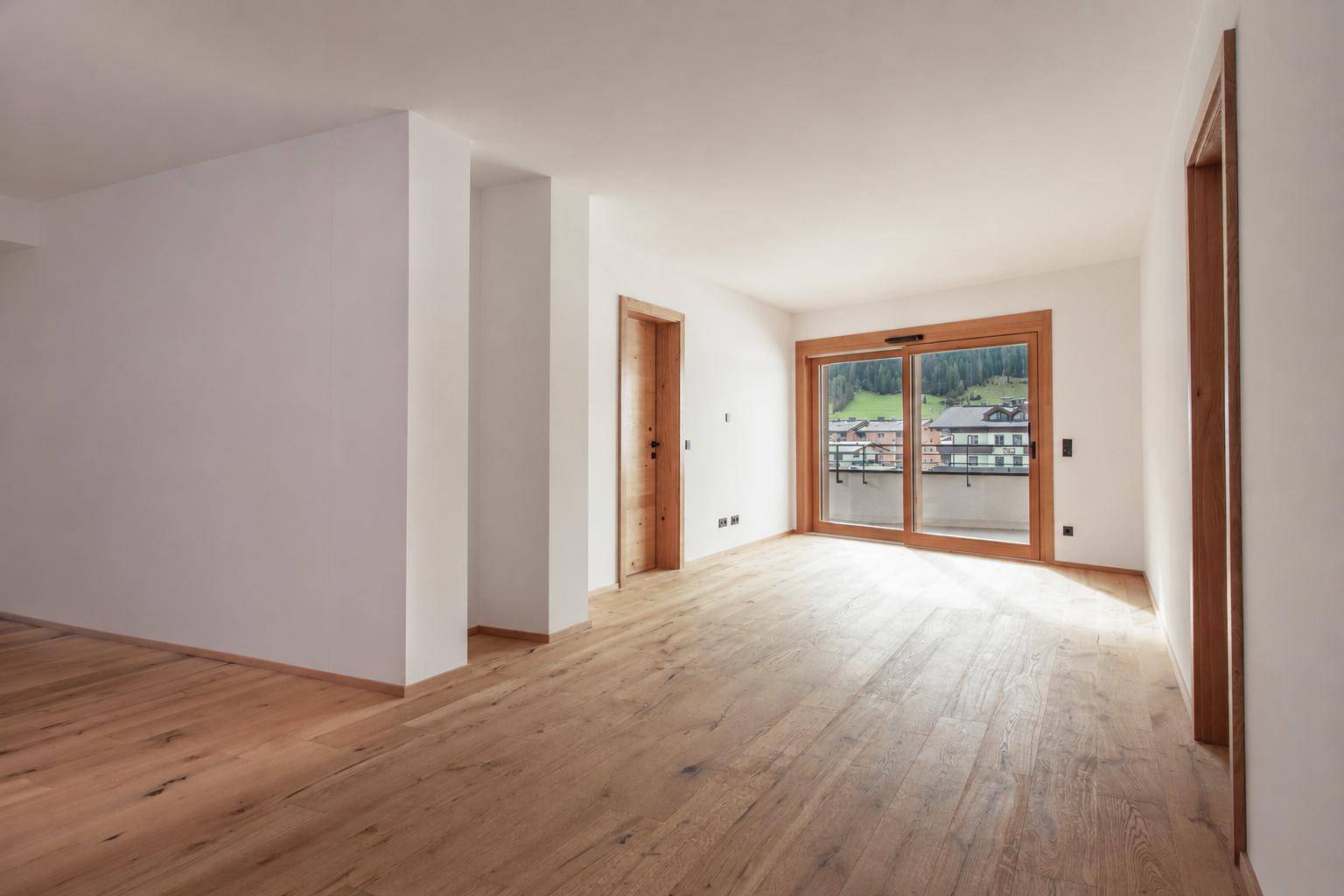 Geräumiger, heller Raum mit Holzboden und Zugang zu einem Balkon mit Blick ins Grüne.