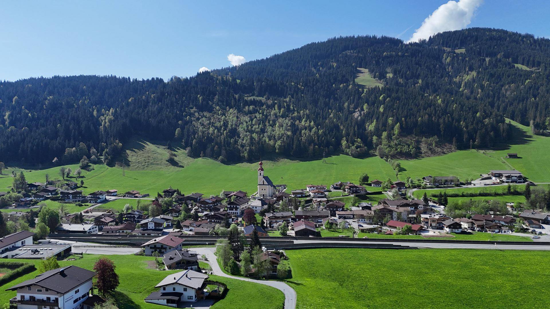 Blick auf ein malerisches Dorf in einem grünen Tal, umgeben von bewaldeten Bergen.