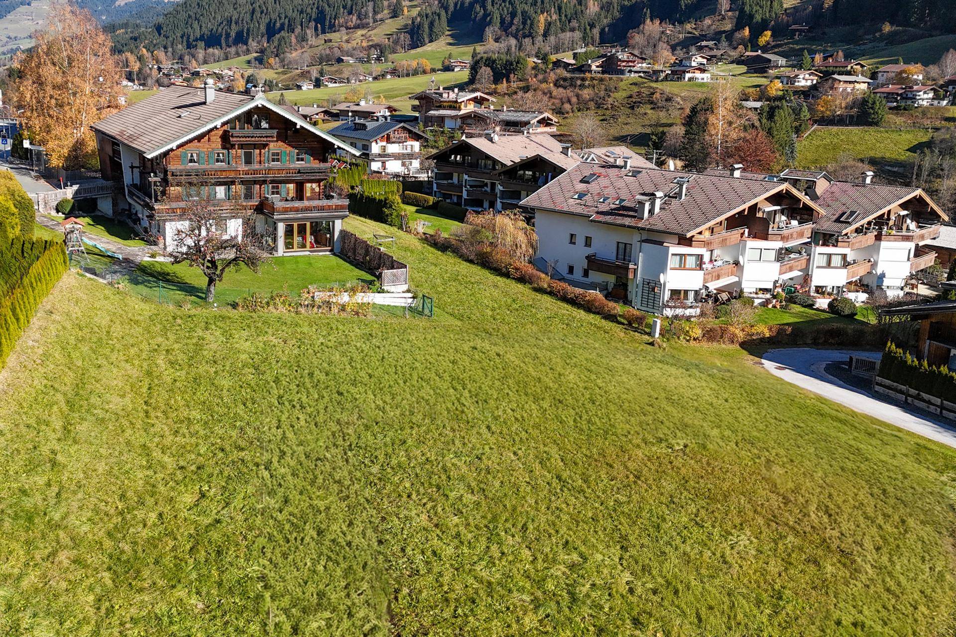 Panoramablick auf mehrere Wohnhäuser am Hang, umgeben von weitläufigen grünen Wiesen und herbstlicher Vegetation.